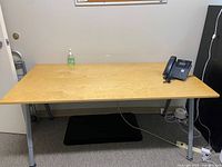 Photo showing the Ikea desk with a light wood grain surface and silver metal legs, with a phone and hand sanitizer on top.