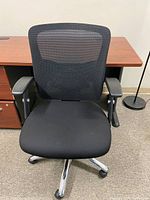 Black office chair with mesh backrest, padded seat, adjustable armrests, and chrome five-wheel base, positioned in an office setting.