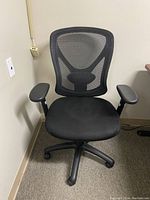 Photo of a black office chair with fabric seat and mesh backrest, adjustable armrests, and five caster wheels.