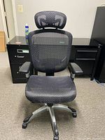 Front facing view of a black mesh ergonomic office chair with adjustable headrest and armrests in office setting.