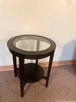 Full view of black round side table with glass top and lower shelf shown against a wall on carpeted floor.