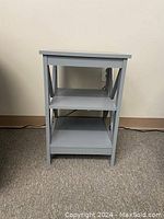 Front view of a light gray wooden side table with two shelves, placed against a white wall on a carpeted floor.