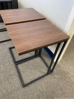 Two rectangular side tables with wood veneer tops and black metal rectangular frame bases placed side by side on carpeted floor near wall.