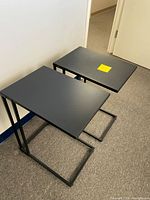 Two black metal-based side tables with rectangular tops placed on a beige carpeted floor near a white wall and door.