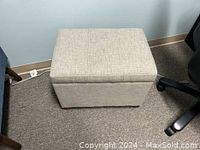Photo showing a gray rectangular fabric storage ottoman with a hinged lid, positioned on a carpeted floor next to a desk chair and power strip.