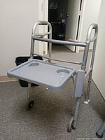 Silver aluminum assistive walker with attached fold-up feeding tray in gray plastic that has two cup holders. Two wheels in front, rubber tips on back legs. Shown standing on hardwood floor in indoor setting.