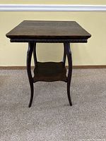 Front view of the antique dark oak side table showing its square top, carved edge details, curved legs, and lower shelf.