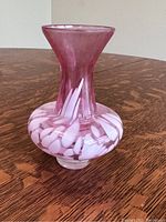 Pink and white art glass vase on wooden surface, showing side profile with narrow neck and flared lip, detailed white swirl patterns around the base.