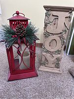 Red Christmas lantern beside tall beige JOY pillar stand on carpeted floor