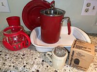 Photo shows white rectangular Corningware baking pan, red Kuhn Rikon pot with lid and handle, red dinner plate, red plastic tumbler, glass microwave popcorn popper with red silicone cover, and Pampered Chef white flour/sugar shaker with box on kitchen countertop.