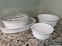 White CorningWare French White oblong pan with clear glass lid and three round casserole dishes on granite countertop.