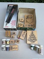 Photo showing Milwaukee Precision Hot Tool, multiple wooden block embossing stamps with seasonal and floral designs, jars of embossing powders and glitter in various colors