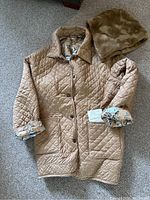 Full view of tan quilted coat laid flat on the floor with sleeves rolled showing floral lining. Detachable faux fur hood placed adjacent.