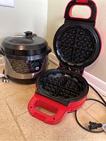 Cooks Essentials pressure cooker with control panel and lid beside Power XL red waffle maker opened showing waffle plates