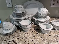 Full set of Noritake Whitehall china including stacked plates, cups, saucers, sugar bowl with lid, cream pitcher, and serving platters on a granite countertop under bright lighting.