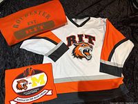 White RIT hockey jersey with orange and black sleeves, tiger logo front and center, folded orange fleece lined stadium blanket with Rochester EST. 1829 RIT print, folded orange souvenir T-shirt with Homecoming event graphics.