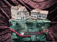 Photo shows two miniature lighted building ornaments, The Antique Shop and Rockwells Residence, sitting on a box labeled Hawthorne Porchlight Collection.