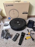 Photo of the Roomba e5 vacuum, charger base, remote control, cleaning brush, accessories, and original box from a different brand (ECOVACS).