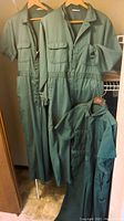 Three vintage green work overalls hanging on hangers, showing two short sleeve and one long sleeve version with zipper fronts and elastic waists.