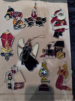Full view of all 9 vintage stained glass Christmas ornaments laid out on a surface. Includes designs of bell, angel, Santa, holly, lantern, train, house, oil lamp, and angel with trumpet. The central angel is thicker in shape than the others.