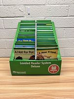 Front view of the green cardboard box with two rows of leveled reader books visible, showing titles like 'A Hat for Pat' and 'A New Friend'.