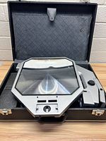 Apollo magnifier set in black case, showing magnifier lens, metal arm with built-in light and adjustment knobs, and interior of the case.