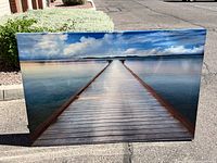 Full view of the large rectangular wall hanging photo print showing the wooden dock leading into calm water with blue sky and clouds above.