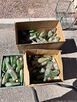 Three containers filled with vintage glass soda bottles, mostly covered in dirt or dust. Many Coca-Cola and Jarritos bottles visible, mostly green and clear glass.