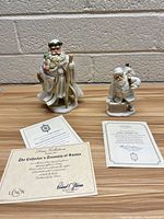 Two Lenox Santa Claus figurines displayed on a wooden surface against a brick background, with certificates of authenticity placed in front.