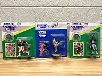 Photo showing all three Starting Lineup figures in original packaging, arranged side by side on a wood surface against a white wall.