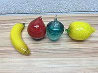 Top-down view of four vintage glass fruit figurines including banana, red pear, teal apple, and lemon, showing color and condition.