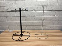 Photo of two metal display racks: a black rack with multiple hooks and circular base, and a white rack with a single hook and circular base, on a wooden surface against a brick wall.