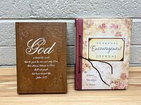 Two books placed side by side on a gray surface against a white brick wall. Brown leather-bound book with John 3:16 quote on the left. Floral-bound journal with title 'Everyday Encouragement Journal' with string clasp on the right.
