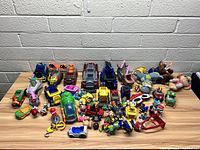 Wide view of assorted Paw Patrol toy figures, plush toys, and vehicles scattered on a wooden surface in front of a light grey wall.