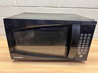 Front angled view of black Sylvania microwave oven on table in front of white wall, showing keypad and windowed door.