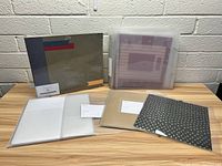 Photo of scrapbooking albums and sleeve protectors arranged on wooden table against white brick wall, showing the front and back covers along with open sleeve protectors