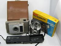 Polaroid J33 Land Camera with attached carrying case, Kodak Tele-Ektralite 600 with flash and original packaging, and Brownie Starflash camera shown together on white background.