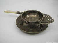 Side angle of metal tea strainer showing perforated bowl, mother of pearl handle, and metal stand beneath.