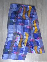 Full view of the scarf laid flat showing the abstract colorful cubist design featuring human faces and geometric shapes in blue, purple, yellow, pink tones.