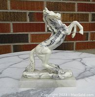 Front and right side view of a white alabaster horse sculpture with black veining in a rearing pose on a rectangular base, placed on a marble surface in front of a brick wall.