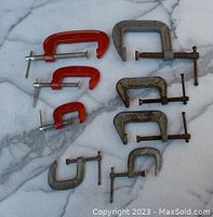 Nine C-clamps arranged on a marble surface, showing variety in size and color (red painted and gray metal).