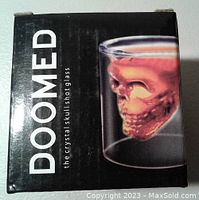 Black box with white text 'DOOMED the crystal skull shot glass' and image of the shot glass filled with red liquid showing skull shape inside.