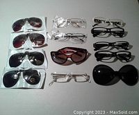 Multiple pairs of eyeglasses and sunglasses arranged on a flat surface, including clear reading glasses, black frames, tortoiseshell sunglasses, and clip-on flip-up sunglasses, all new.