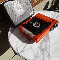 Maris portable orange gas stove with black burner and control knob, and black plastic carry case, all displayed on a white marble table.