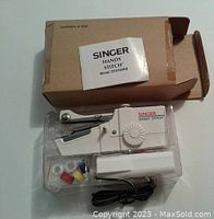 Singer Handy Stitch handheld sewing machine in plastic tray with power cord, multiple thread spools, instruction manual, and original box.