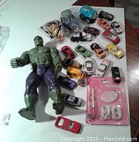 Photo showing large green Hulk action figure, numerous small toy cars in various colors, blue cartoon animal face vehicle toy, black toy train engine, and birthday candle set.