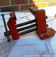 Side and angled view of the red vise showing screw handle and jaws, placed on a white marble table.