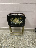 Set of four folded vintage TV trays showing black top surfaces with floral painting and gold-tone metal legs with wooden handles.