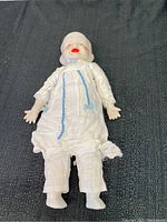 Porcelain doll full body lying on black textured fabric surface, dressed in white clothes with blue accents.