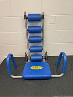 Front view of the Ab Rocket abdominal exercise equipment showing blue seat, padded rollers, and curved handles.